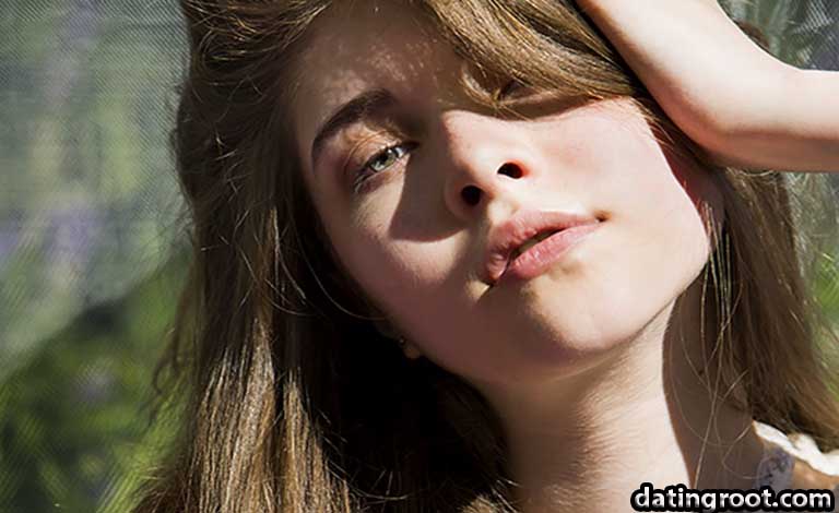 Close-up portrait of a young woman outdoors, hand resting on her head, sunlit skin and soft expression, blurred greenery in the background.