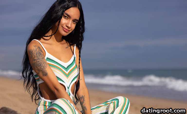 Woman with long dark hair sitting on a sandy beach, wearing a green, white, and orange striped outfit, tattoos on her arms with ocean waves in the background